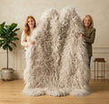 Two women holding up a large, fluffy shaggy faux fur throw blanket in a warm neutral-toned room with a potted plant and wooden floor.