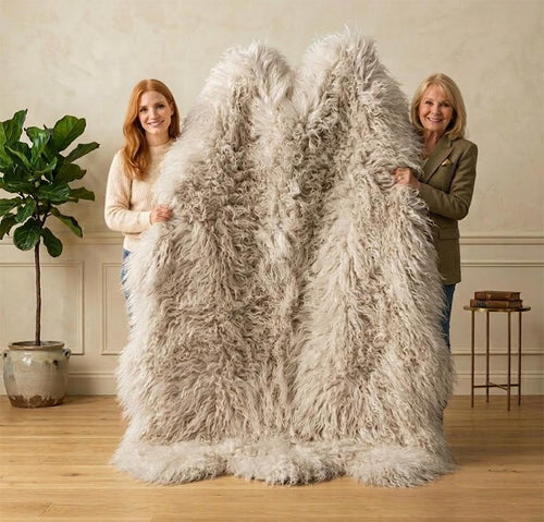Two women holding up a large, fluffy shaggy faux fur throw blanket in a warm neutral-toned room with a potted plant and wooden floor.